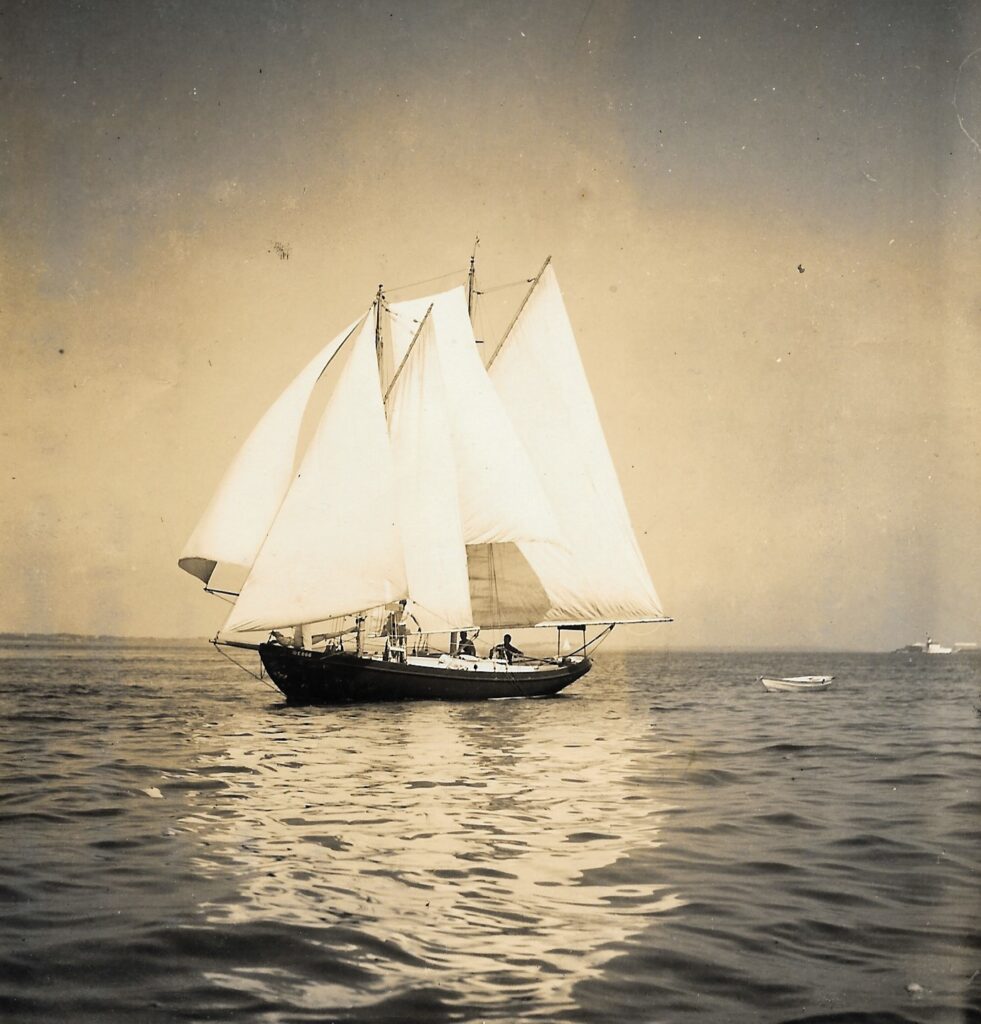 Arnie Gay's schooner Delilah with two people aboard. Delilah's sails are filled with a gentle breeze and a small skiff is in tow.