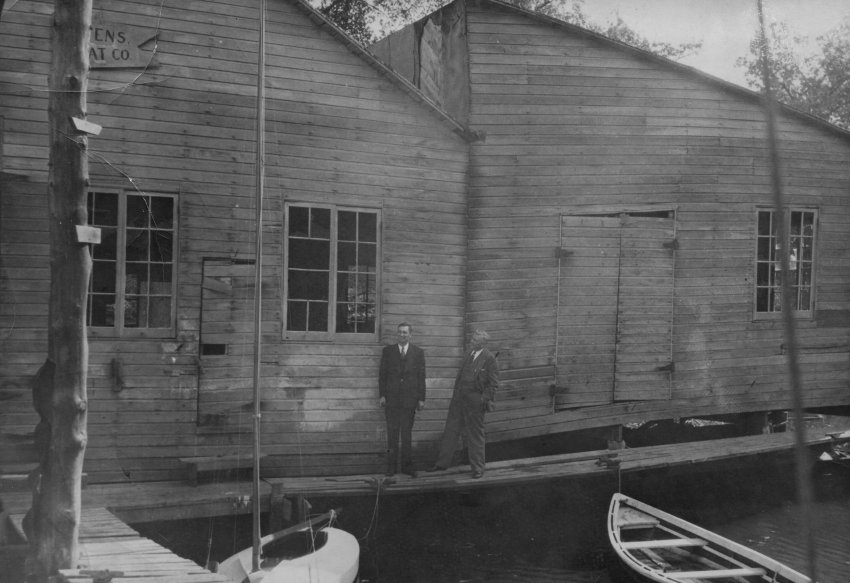 In this photograph, taken about 1941, two unidentified men in suits can be seen standing on a narrow, wooden walkway outside the Owens Boat Company buildings. Part of the Owens Boat Company sign can be seen in the upper right corner. Also seen in the picture are a wooden dock, a small sailboat, and a small skiff.