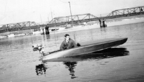 Pictured here is a young Norman Owens in a miniature hydroplane boat with an outboard motor on Spa Creek in 1929. In the background a bridge and several docked boats can be seen.