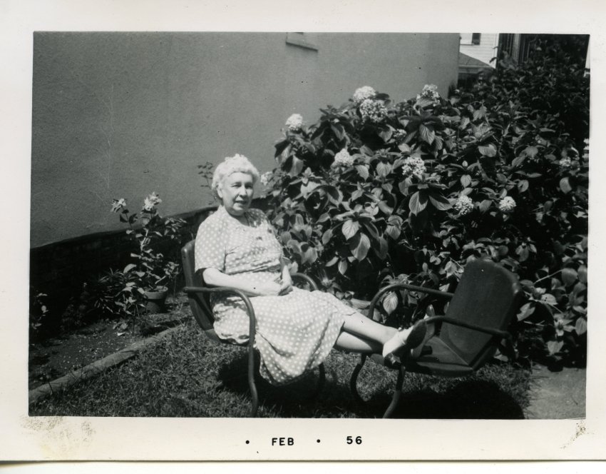 Pearl McNasby seated in a chair with her feet resting on another chair. A large flower bush blooms behind her. A date is stamped along the bottom of the photo, "Feb '56".